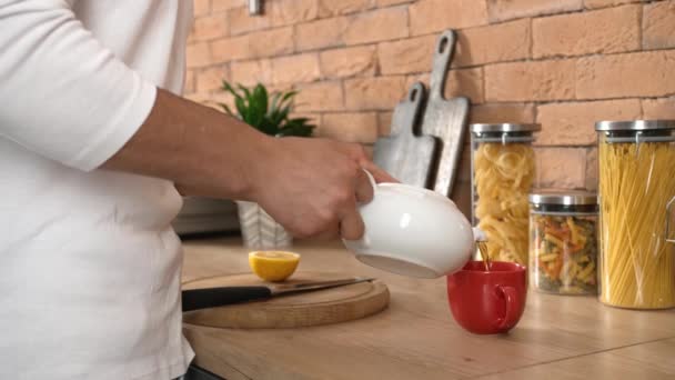 若い男がティーポットからカップに熱いお茶を注ぎ 自宅で台所でレモンを切断 — ストック動画