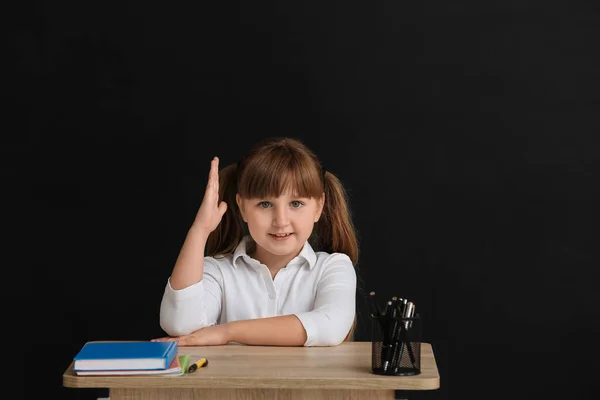 Little pupil with raised hand sitting at school desk against dark background