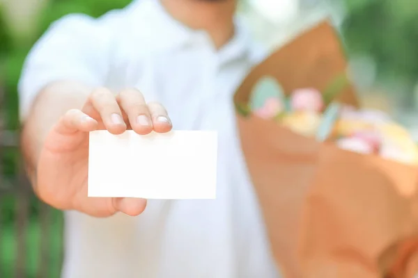 Delivery man with bouquet of beautiful flowers and blank business card outdoors, closeup
