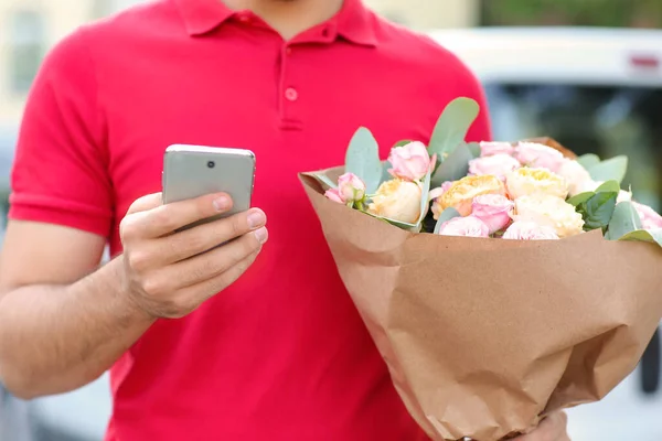 Delivery man with bouquet of beautiful flowers and mobile phone outdoors, closeup