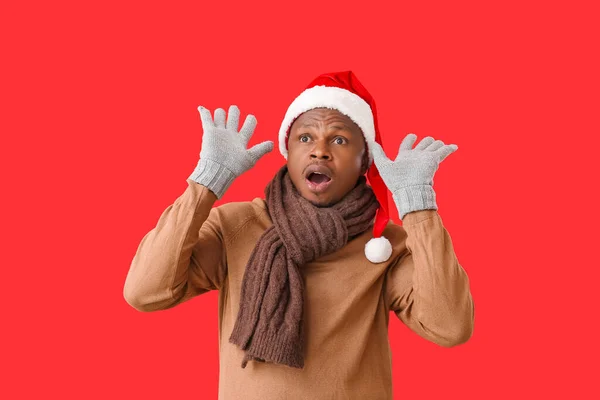 Shocked African-American man in Santa hat and gloves on color background