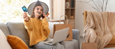 Cadı şapkalı genç bir kadın Cadılar Bayramı için internette kredi kartı alışverişi yapıyor.