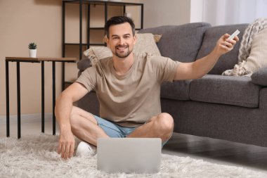 Handsome young man switching on air conditioner while using laptop in living room