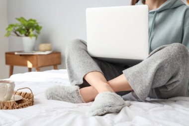 Young woman in soft slippers resting on bed with modern laptop