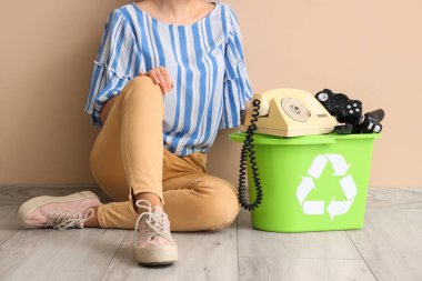 Bej duvarın yanında elektronik aletleri olan geri dönüşüm kutusuyla oturan kadın. Geri dönüşüm kavramı