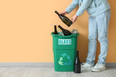 Bej duvarın yanında GLASS tabelası olan bir kadın şişeyi çöp kutusuna koyuyor. Geri dönüşüm kavramı