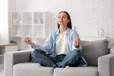 Evdeki kanepede meditasyon yapan genç ve güzel bir kadın.