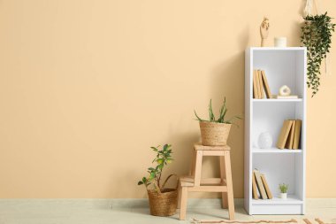 Kitaplık ve tabure. Odada bej duvarın yanında bitkiler var.