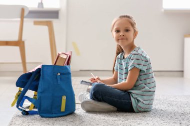 Cute little girl with backpack, notebook and pencil sitting on floor and studying at home