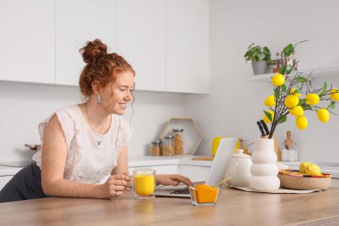 Mutfaktaki masada dizüstü bilgisayar kullanan, zerdeçal içen kızıl saçlı genç bir kadın.