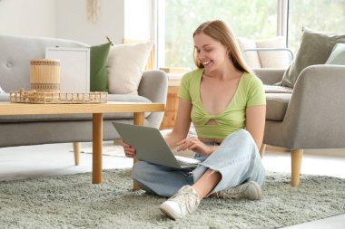 Genç ve güzel bir kadın şık bir oturma odasında dizüstü bilgisayar kullanıyor.