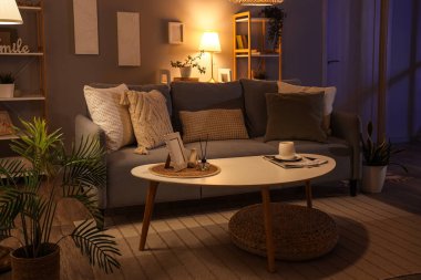 Interior of dark living room with glowing lamps, sofa and shelf units in evening