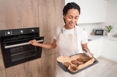 Young African-American woman taking baking dish with tasty cookies from oven in kitchen