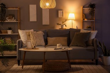 Interior of dark living room with glowing lamps, sofa and shelf units in evening