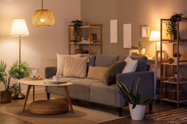 Interior of dark living room with glowing lamps, sofa and shelf units in evening