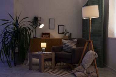 Interior of dark living room with glowing lamps, armchair and table in evening