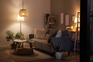 Interior of dark living room with glowing lamps, sofa and plants in evening