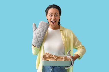 Happy young African-American woman holding baking dish with tasty cookies on blue background