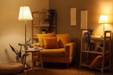 Interior of dark living room with glowing lamps, armchair and shelf units in evening