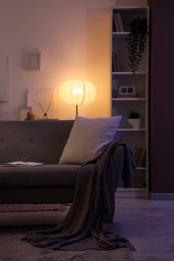 Interior of dark living room with glowing lamp, sofa and shelf unit in evening