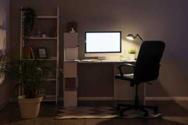 Interior of dark office with glowing lamp, workplace and shelf unit in evening