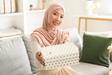 Salondaki kanepede oturan hediye kutusuyla tesettürlü güzel Asyalı kadın.
