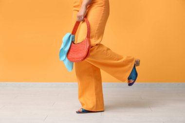 Sarı duvarın yanında turuncu çantalı, pantolonlu, topuklu ayakkabılı şık genç bir kadın.