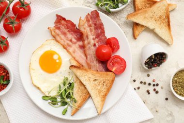 Plate with tasty fried egg, bacon, toasts, tomatoes and spices in bowls on white grunge background. Closeup