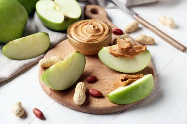 Cutting board with fresh apple slices and bowl of sweet peanut butter on white wooden background