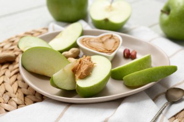 Plate with fresh apple slices and bowl of sweet peanut butter on white wooden background, closeup