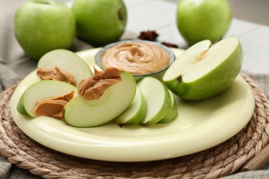 Plate with cut fresh apple and bowl of sweet peanut butter on white wooden background, closeup
