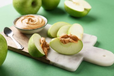 Cutting board with fresh apple slices and bowl of sweet peanut butter on green background, closeup