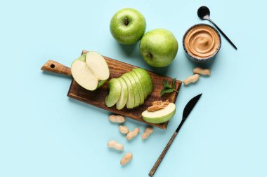 Wooden board with fresh apple slices and bowl of sweet peanut butter on blue background