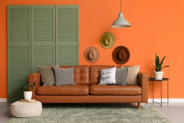 Brown leather sofa with soft pillows and folding screen near orange wall in living room