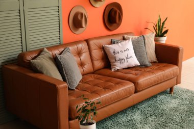 Brown leather sofa with soft pillows and folding screen near orange wall in living room