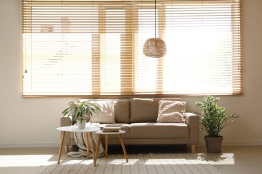 Oturma odasının penceresinin yanında masaları ve çiçekleri olan rahat kahverengi bir kanepe.