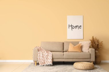 Oturma odasındaki bej duvarın yanında hasır pouf ve pampa otlu rahat kahverengi kanepe.