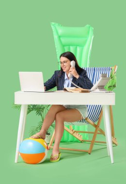 Laptoplu genç iş kadını yeşil arka plandaki masada güverte sandalyesinde telefonla konuşuyor. Yaz tatili kavramı