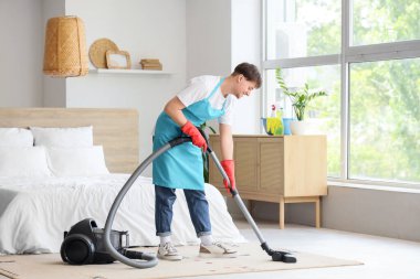 Young male janitor with modern vacuum cleaner cleaning carpet in bedroom