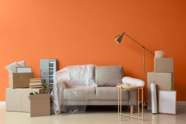 Grey sofa with stretch film and boxes near orange wall in room on moving day
