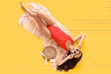 Sarı arka planda duran plaj aksesuarları ve güneş kremi ile kırmızı mayo giymiş güzel, mutlu bir Asyalı kadın.