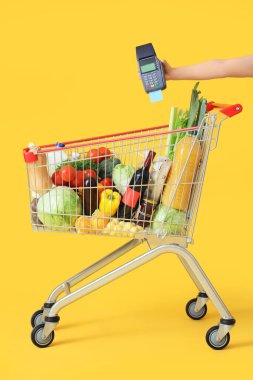 Female hand holding payment terminal and shopping cart with different fresh products on yellow background