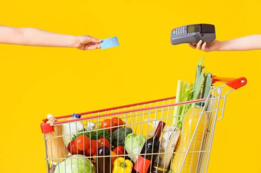 Female hands with credit card, payment terminal and shopping cart full of different fresh products on yellow background