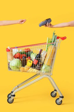Female hands with credit card, payment terminal and shopping cart full of different fresh products on yellow background