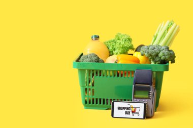 Yeşil alışveriş sepeti, farklı yeni ürünler, ödeme terminali ve sarı arka planda cep telefonu.