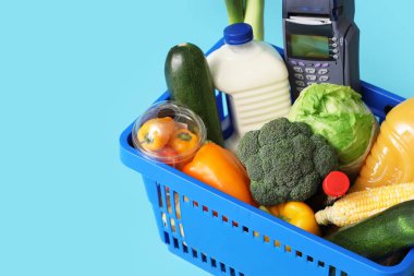 Shopping basket with different fresh products and payment terminal on blue background, closeup