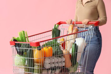 Woman with shopping cart full of different fresh products on pink background