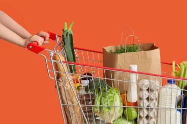 Female hands with shopping cart full of different fresh products on orange background