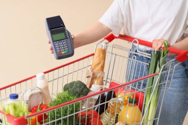 Woman with payment terminal and shopping cart full of different fresh products on beige background