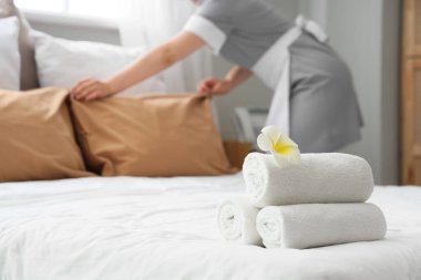 Young female chambermaid with clean towels and plumeria flower making bed in hotel room, closeup